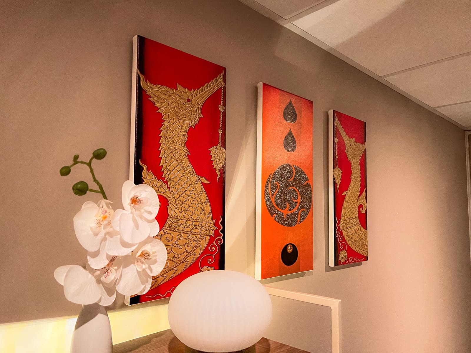 Thaise schilderijen in rood en goud boven een sidetable met witte orchideeën